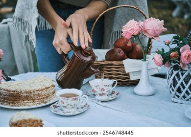 Woman pouring tea at an outdoor picnic with cake and roses. Enjoy the cozy autumn atmosphere. Perfect for blogs, websites, or social media. - Powered by Shutterstock