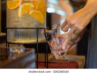 Woman Pouring Water From Dispenser. Detox Fruit Infused Flavored Water, Lemonade In A Beverage Dispenser With Orange Juice, Healthy Drink For Lifestyle.