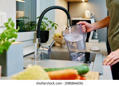 Woman pouring water from faucet into water filter jug at the kitchen
