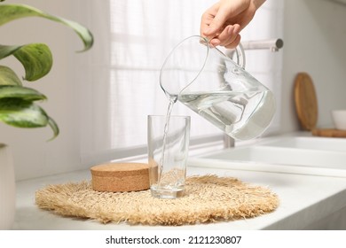 Woman Pouring Water Into Glass From Jug On Countertop In Kitchen, Closeup