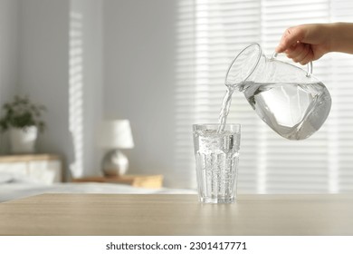Woman pouring water from jug into glass at wooden table indoors, closeup. Space for text