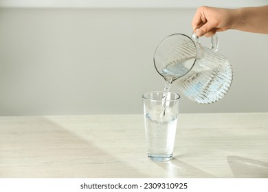 Woman pouring water from jug into glass on white table indoors, closeup. Space for text