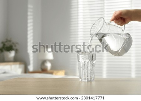 Woman pouring water from jug into glass at wooden table indoors, closeup. Space for text