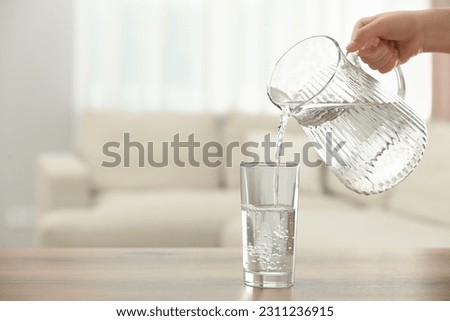 Woman pouring water from jug into glass at table indoors, closeup. Space for text