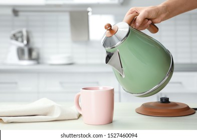 Woman pouring water from modern electric kettle into cup at wooden table in kitchen, closeup
