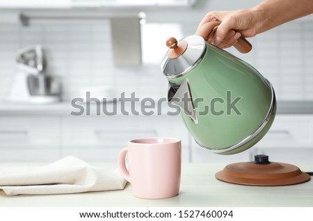 Woman pouring water from modern electric kettle into cup at wooden table in kitchen, closeup