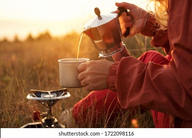 Woman Is Pours Coffee From Cup With Coffee Pot On Background Of Sunrise Meadow In Mountains. Tourist Hiker Makes Transition, Meets Dawn Sun. Lens Flare. Camping. Lifestyle. Travel