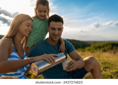 A woman pours coffee from a thermos to her husband, while a girl smiles and stands behind them - Powered by Shutterstock