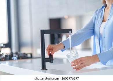 Woman Pours Fresh Filtered Purified Water From A Tap Into A Glass At Kitchen At Home For Drinking 