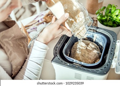 Woman Pours Ingredients Into The Bread Maker Machine. Photo Taken In The Light Of Natural Sun And Studio Lamps. It Contains Delicate Artistic Noise And A Vintage Color Grading