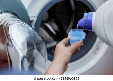 Woman pours liquid washing gel into plastic cap against background of drum of steel-colored washing machine. A girl carefully pours a transparent conditioner for flattening laundry