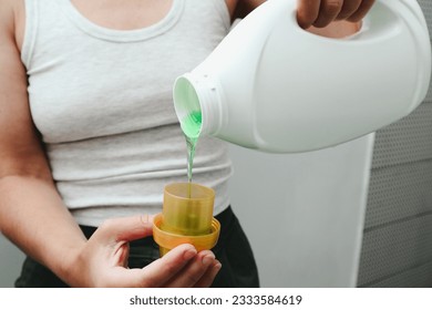 Woman pours liquid washing gel into plastic cap. Concept of washing clothes, domestic routine and household chores. Close up of attractive female pouring liquid green gel into bottle cap. - Shutterstock ID 2333584619