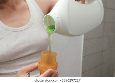 Woman pours liquid washing gel into plastic cap. Concept of washing clothes, domestic routine and household chores. Close up of attractive female pouring liquid green gel into bottle cap. - Shutterstock ID 2336297227