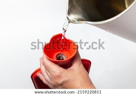 a woman pours a red rubber hot-water bottle with boiling water from a teapot