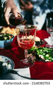 A Woman Pours Red Wine Into A Glass On The Table. Dinner With A Large Group Of Friends In The Countryside. Vertical Photo.