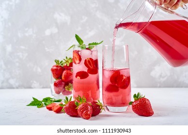 Woman Pours Strawberry Lemonade In Glass. Cocktail With Strawberry, Ice And Mint In Glasses On White Concrete Background, Copy Space. Refreshing Summer Berry Drink