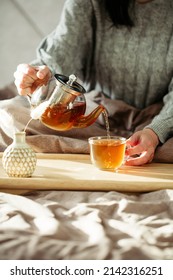 Woman pours tea into glass mug from teapot, she sitting in bed with wooden tray. Breakfast in cozy home room interior. Nordic, hygge bedroom.