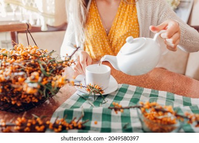 Woman Pours Tea With Sea Buckthorn Berries In Cup From Kettle. Healthy Hot Drink For Cold Fall And Winter Days. Yellow Fruit On Harvested Branches