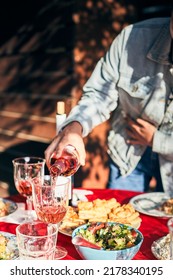 A Woman Pours Wine Into Glasses. A Full Table Is Set For A Friendly Banquet Outside. Vertical Photo.