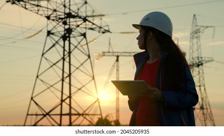 A Woman Power Engineer In A Safety Helmet Checks The Power Line Using A Digital Tablet. Electricity Company Employee Woman Working Outdoors, Servicing High Voltage Electrical Lines At Sunset