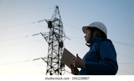 A Woman Power Engineer In White Helmet Inspects Power Line Using Data From Electrical Sensors On A Tablet. High Voltage Electrical Lines At Sunset. Distribution And Supply Of Electricity. Clean Energy