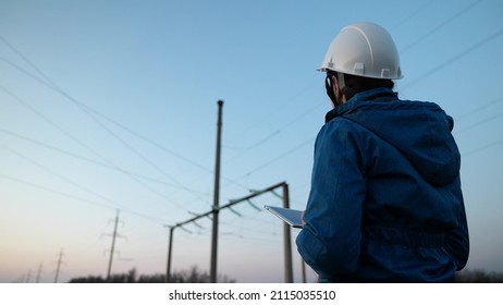 Woman Power Engineer In White Helmet Checks Power Line Using Computer On Tablet, Remote Control Of Power System. High Voltage Electrical Lines At Sunset. Distribution And Supply Of Electricity. Energy