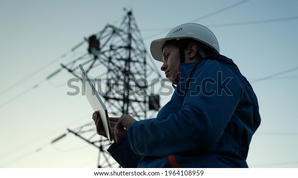 Woman power engineer in white helmet checks power\
line using computer on tablet, remote control of power system. High\
voltage electrical lines at sunset. Distribution and supply of\
electricity. Energy