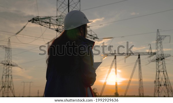 Woman power engineer in a white helmet checks\
power line using data from electrical sensors on a tablet. High\
voltage electrical lines at sunset. Distribution and supply of\
electricity. clean energy