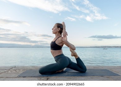 A woman practices the Eka Pada Rajakapotasana yoga pose on the river bank