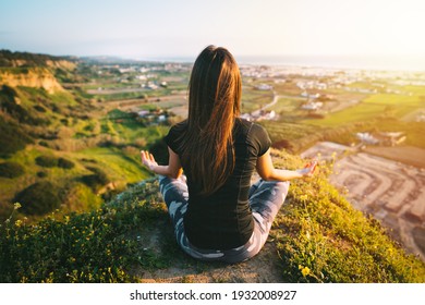 Woman practices yoga and meditates on the top of the mountain with beautiful view on the city and ocean at sunset. Woman sitting in easy pose or sukhasana with mudra. Relaxation, harmony with nature