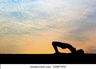 Woman practicing bridge yoga pose outdoors over sunset sky background.