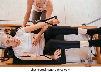Woman practicing stretching exercise with personal trainer on reformer. Top view of elderly grey hair female client maintaining her fitness while exercising on reformer machine in pilates studio.