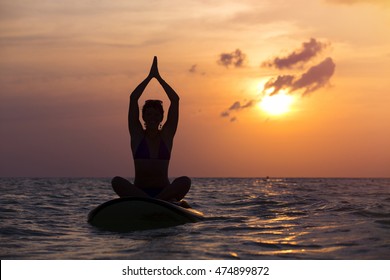 Woman practicing SUP yoga at sunset, meditating on a paddle board.