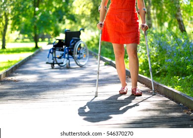 woman practicing walking on crutches in green park and wheelchair standing in the background 