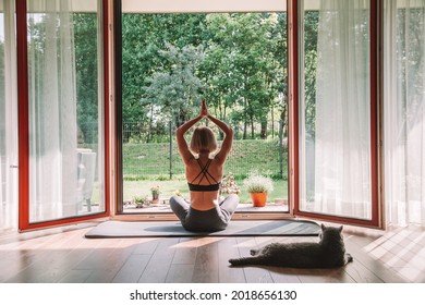 Woman Practicing Yoga At Home, Sitting In A Lotus Position In Front Of The Opened Window. Her Cat Is Watching Her, Laying Down Next To Her.