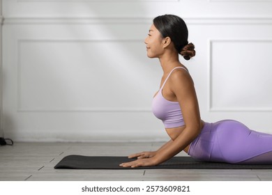A woman practicing yoga indoors, performing cobra pose on yoga mat, wearing purple workout outfit, with calm and focused expression in bright and minimalistic room