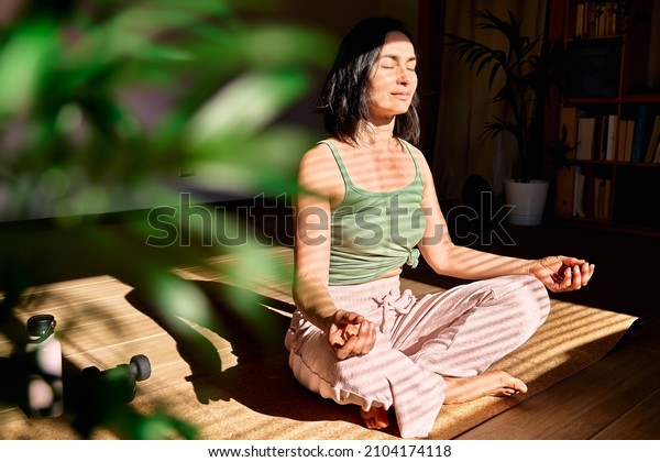 Woman practicing yoga and meditation at home\
sitting in lotus pose on yoga mat, relaxed with closed eyes.\
Mindful meditation concept.\
Wellbeing.
