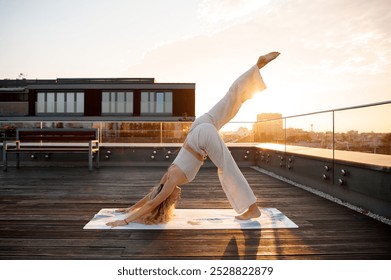 A woman practicing yoga on a rooftop at sunset, performing a downward dog pose with one leg raised. She is wearing a white outfit and is on a yoga mat, with a cityscape in the background. - Powered by Shutterstock