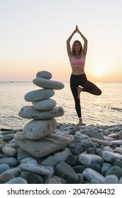 A Woman Practicing Yoga, On The Seashore In The Evening Performs The Vrikshasana Exercise, A Tree Pose, A Balancing Cairn Of Stones Stands Nearby In Defocus