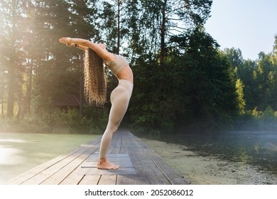 A woman practicing yoga performs an exercise, back bend, Hasta Uttanasana pose, stands on a wooden bridge on a sunny warm morning in a park near a pond