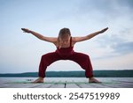 Woman practicing yoga in a wide stance on a wooden pier near a lake. Performing Goddess Pose Utkata Konasana with arms extended, dressed in a red outfit