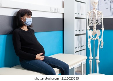 Woman With Pregnancy Belly Wearing Face Mask At Checkup Appointment In Cabinet, Waiting On Doctor To Do Medical Consultation. Patient Expecting Baby Sitting In Office For Health Care Examination