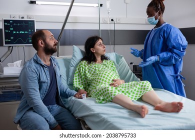 Woman With Pregnancy Talking To Obstetrics Nurse While Sitting In Hospital Ward Bed, Holding Hand Of Husband. Couple Waiting For Baby Delivery And Childbirth At Medical Facility