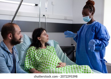 Woman With Pregnancy Talking To Obstetrics Nurse While Sitting In Hospital Ward Bed, Holding Hand Of Husband. Couple Waiting For Baby Delivery And Childbirth At Medical Facility