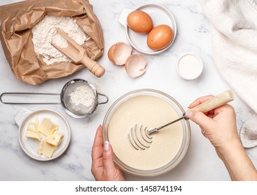 Woman prepares dough for homemade pancakes for Breakfast. Whisk for whipping in hands. Ingredients on the table - wheat flour, eggs, butter, sugar, salt, milk. Selective focus