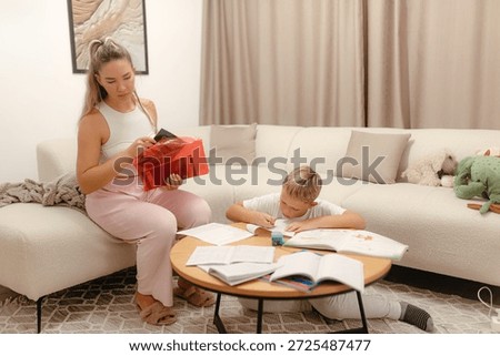 Woman prepares study folder while boy finishes workbook task near low table, warm beige room, peaceful focused atmosphere.