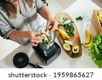Woman is preparing a healthy detox drink in a blender - a  green smoothie with fresh fruits, green spinach and avocado. Healthy eating concept, ingredients for smoothies on the table, top view