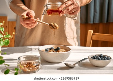 Woman preparing healthy dieting vegan nutritious breakfast. Female hand pouring honey in the bowl with oatmeal porridge with walnuts and blueberries. - Powered by Shutterstock