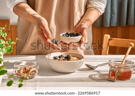 Woman preparing healthy dieting vegan nutritious breakfast. Female hand putting blueberries in the bowl with oatmeal porridge with walnuts and honey.