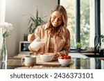 A woman preparing herself breakfast, pouring milk into a plate with cereal, strawberries and a cup of coffee standing nearby. A woman is having breakfast in a cozy kitchen.